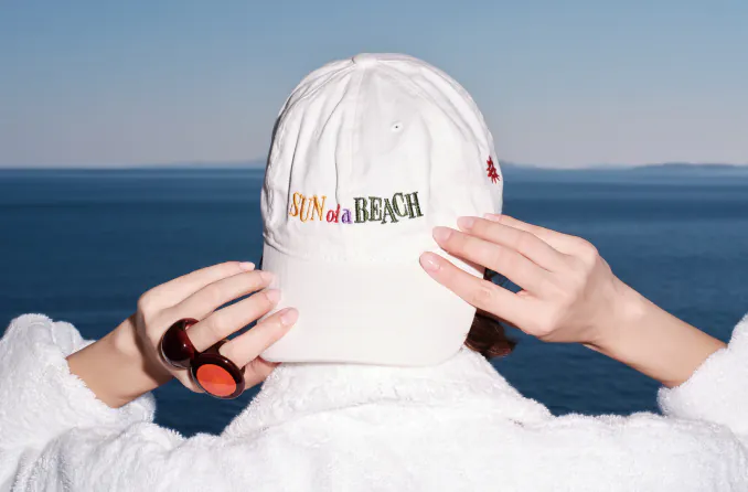 A woman wearing a 'Sun of a Beach' hat facing the sea, showcasing a relaxed and stylish vacation vibe.